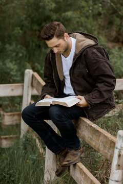 A young man reads from his Bible as he sits on a rail fence in the countryside; Alberta, Canada