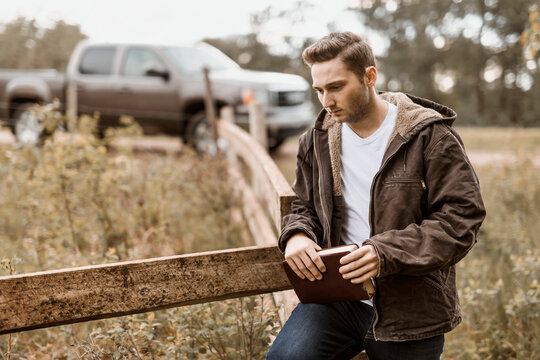 A young man stands holding his Bible beside a rail fence and truck park on a road in the countryside; Alberta, Canada