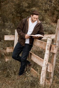 A young man reads from his Bible as he stands against a rail fence in the countryside; Alberta, Canada