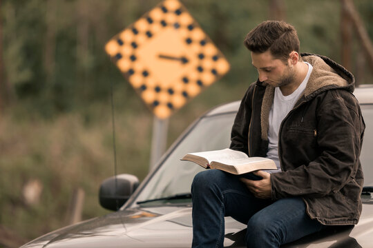 A young man reads from his Bible as he sits on a pickup truck hood in the countryside, a directional roadside sign with arrow blurred in the background; Alberta, Canada