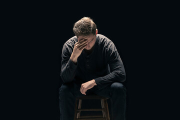 A young man sits with his face in his hands against a black background; Studio