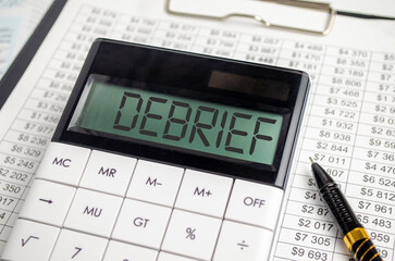 On a wooden table there is a black pen and a calculator with the text DEBRIEF. Business concept