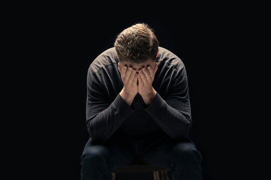 A young man sits with his face in his hands against a black background; Studio