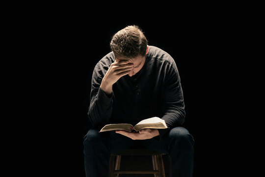 A young man sits with his hand holding his head as he looks down reading his Bible against a black background; Studio