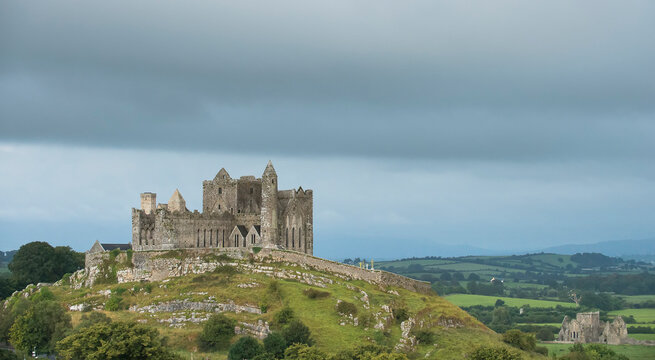 The ancient Rock of Cashel on the hilltop against a cloudy sky with Hore Abbey and the Tipperary countryside below; Cashel, County Tipperary, Ireland