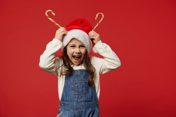 Mischievous happy little girl holding Christmas stripped lollipops on her head as horns, rejoicing at upcoming winter holidays, smiling at camera, isolated on red background, copy advertising space