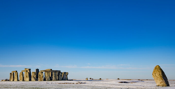 Stonehenge defined by early morning snow with a bright blue sky and the leaning Heel Stone off to the right of the stone circle; Wiltshire, England, United Kingdom