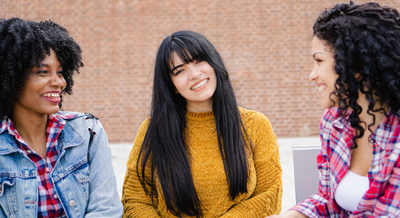 Fototapeta premium Three young, Latina and Hispanic interracial college friends chatting on a campus bench between classes.