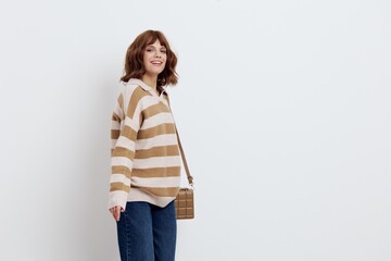 a pretty sweet woman is standing on a light background in a striped sweater and a bag on her shoulder, standing straight with her shoulders slightly twisted