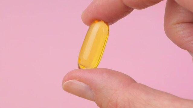 Woman holding a yellow Vitamin D or fish oil gel Capsule in hand on pink background, omega-3 daily supplement in golden pills, close up slow motion video. High quality horizontal 4k footage