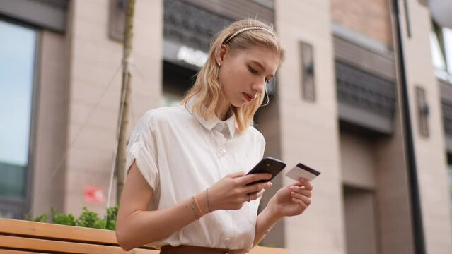 Low-angle view of smiling female tourist holding bank card, entering cvv code or payment information in mobile internet shopping application, sitting on bench on city street. Shooting in slow motion.