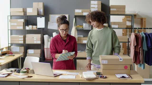 Female manager using laptop and taking notes while giving parcels to young male courier during workday in delivery service office