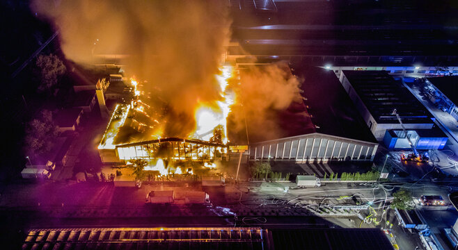 Big fire of an industrial company in the night taken by a drone. Firefighters in action to extinguish a large fire within an industrial area
