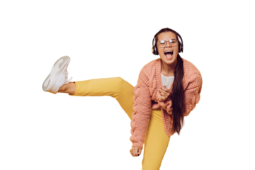 Young cheerful brunette in glasses dancing, listening music in headphones high lifting her leg, in yellow pants, white shoes, pink sweater and headphones on her head over transparent background.