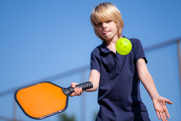 Happy blonde boy playing pickleball game, hitting pickleball yellow ball with paddle, outdoor sport...