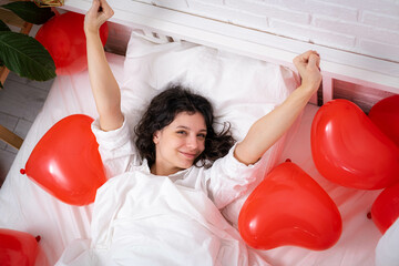 Brunette European girl wake up on bed with red heart shape balloons. Morning love surprise gift on...