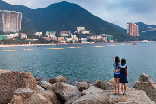Young sisters stand in an embrace looking out at Repulse Bay in Hong Kong; Hong Kong, China