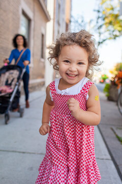 Preschooler girl running down a sidewalk in her red and white gingham dress and looking into the camera with her mother and a stroller in the background; Toronto, Ontario, Canada