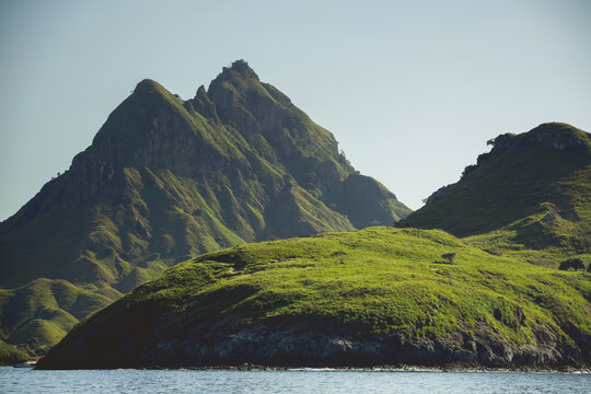 Rugged landforms covered in green foliage along the ocean coastline under a blue sky, Komodo National Park; East Nusa Tenggara, Indonesia