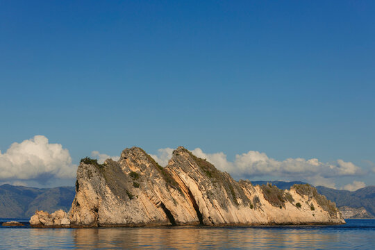Rugged landforms along the ocean coastline under a blue sky with cloud resting over mountaintops, Komodo National Park; East Nusa Tenggara, Indonesia