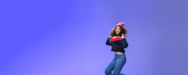 Fototapeta na wymiar Studio shot of cute girl with curly hair in beanie sweatshirt and sneakers jumping playful and carefree over blue background, having fun enjoying cool weather smiling broadly as flying in air