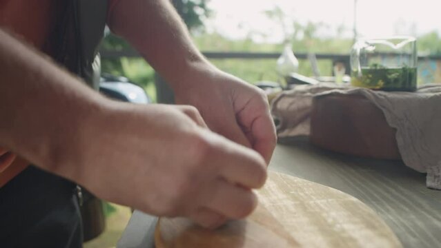 Close up shot of hands on male chef crushing garlic clove with knife on wooden cutting board while preparing food on outdoor terrace