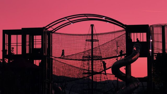 Silhouettes of kids playing on the outdoor park playground against pink sunset sky