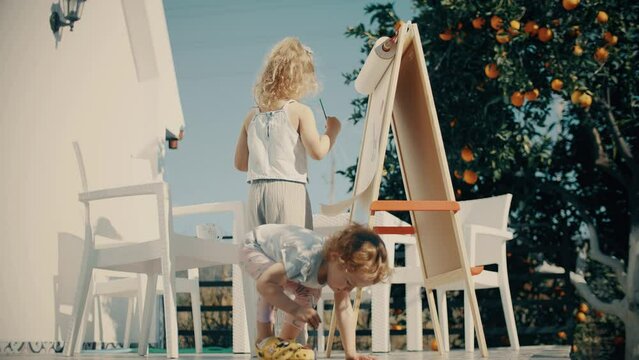 Two adorable little girls paint outdoor at the orange tree on a sunny summer day