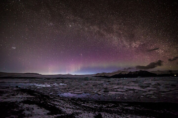 Fototapeta na wymiar Aurora boreale all'orizzonte sormontata dalla via lattea da sinistra a destra con la laguna ghiacciata in primo piano e le montagne sullo sfondo