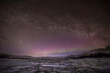 Aurora boreale all'orizzonte sormontata dalla via lattea con la laguna ghiacciata in primo piano e le montagne sullo sfondo