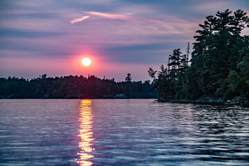Vibrant sun glowing over tranquil lake and forest at sunset and sunbeam reflected on water, Lake of the Woods, Ontario; Kenora, Ontario, Canada