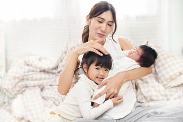 Obraz na płótnie Canvas Asian mother and children girl baby together at home. Asia mom holdind infant and play with her sister sitting on bed in living room at house family having fun together.