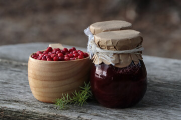 Tasty lingonberry jam in jars and cup with red berries on wooden table outdoors - obrazy, fototapety, plakaty