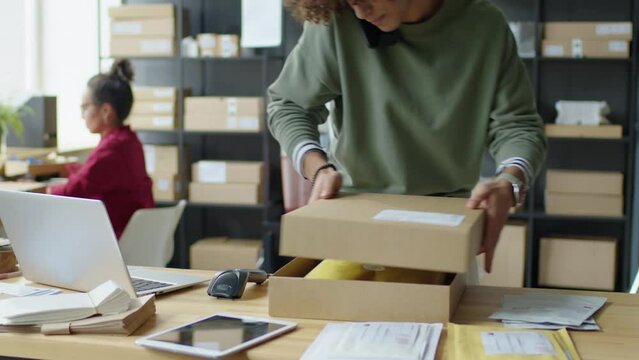 Male worker speaking on phone, packing parcel and scanning label on box while preparing customer order for shipping