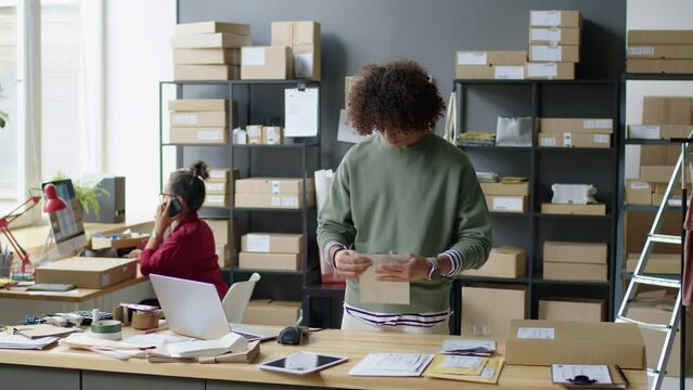Male worker packing parcel in paper envelope, scanning barcode and using laptop while preparing order for express shipping in delivery service office