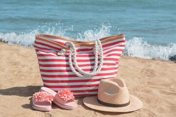 Obraz premium Stylish striped bag with straw hat and slippers on sandy beach near sea
