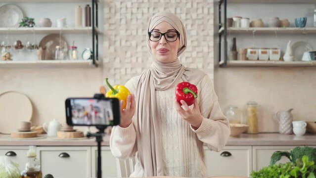 Young muslim woman blogger holding red and yellow pepper live streaming and recording video recipe for food blog. Young pretty female in hijab cooking food online at home kitchen.