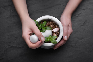 Woman mixing peppercorns, basil and garlic in mortar at black table, top view