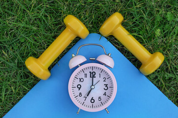 Alarm clock, dumbbells and fitness mat on green grass, flat lay. Morning exercise