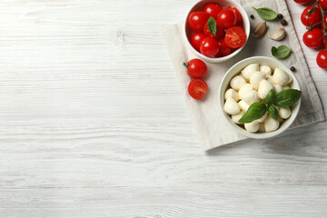 Delicious mozzarella balls in bowl, tomatoes and garlic on white wooden table, flat lay. Space for text