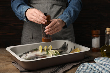 Woman grinding pepper onto raw sea bass fish with lemon and rosemary at wooden table, closeup