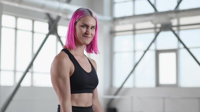 Portrait of a girl performing strength workout while training alone in bright gym studio. Cheerful female athlete smiling while doing dumbbell exercises. High quality 4k footage