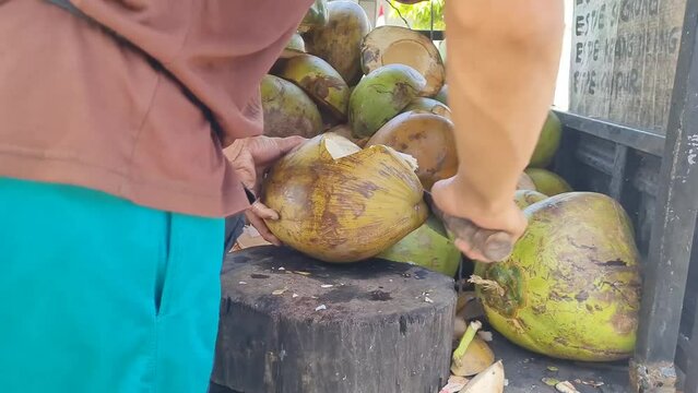 close-up of the hands of an Indonesian iced coconut seller cutting coconuts using a large knife. Street fruit seller's hands peel fresh green coconuts with the help of a cutting knife