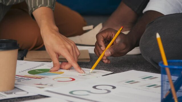 Medium close-up of unrecognizable ethnically diverse man and woman sitting on floor working on business project using graphs and charts