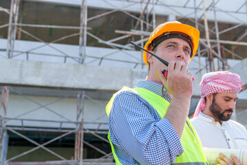 American man technician Industrial engineer using walkie-talkie and holding blueprint working in construction site for building site survey in civil engineering project.