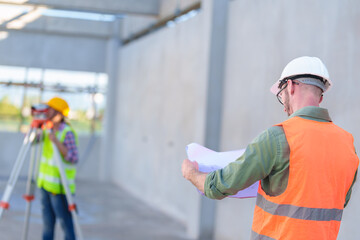 Construction Worker Using Theodolite Surveying Optical Instrument for Measuring Angles in Horizontal and Vertical Planes on Construction Site. Engineer and Architect Using blueprint Next to Surveyor.