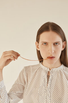 Attractive gender neutral model with long hair pulling soft chewing gum from mouth and looking at camera with serious face expression while standing on gray background
