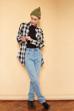 Lovely gender neutral model in unisex outfit buttoning sleeve of checkered shirt while leaning on brown wall