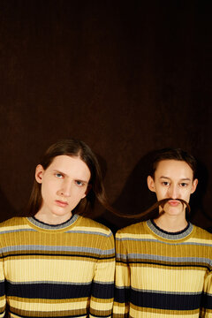 Pretty androgynous female in striped dress making moustache from hair of handsome male in same outfit while standing against brown background together