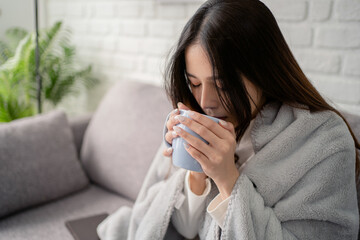 Young woman wearing sweater sitting at home on sofa wrapped in blanket She is drinking hot coffee or tea.